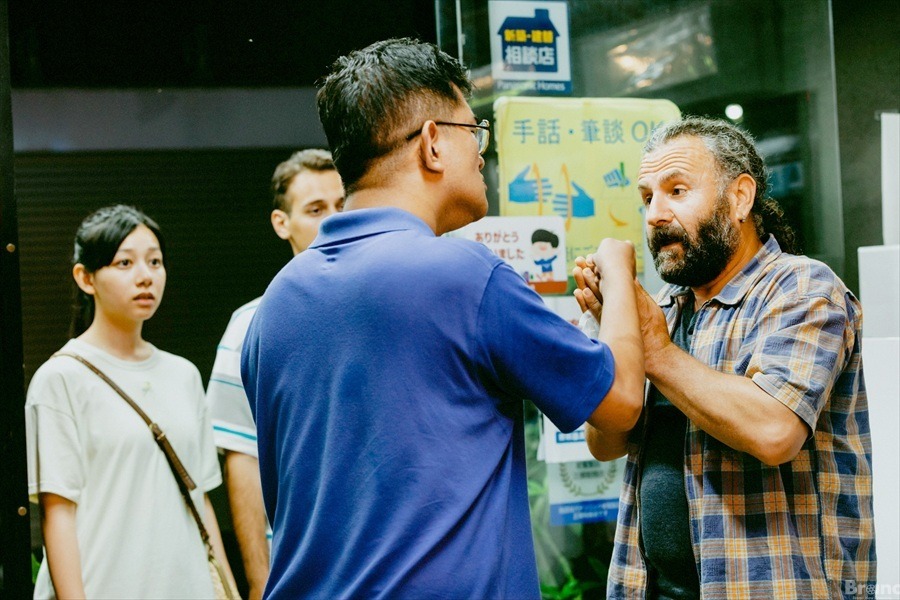 映画『みんな、おしゃべり！』の1場面。店先で、チェック柄のシャツを着た髭のクルド人男性と、青いポロシャツを着た眼鏡をかけた、ろう者の男性が、真剣な表情で向き合い、何かを言い争っている。その様子を、左隣にいる長い髪の女性が心配そうな表情で見つめている。背景のガラス戸には「手話・筆談 OK」と書かれた黄色いステッカーや、「Panasonic Homes」の看板が貼られている。