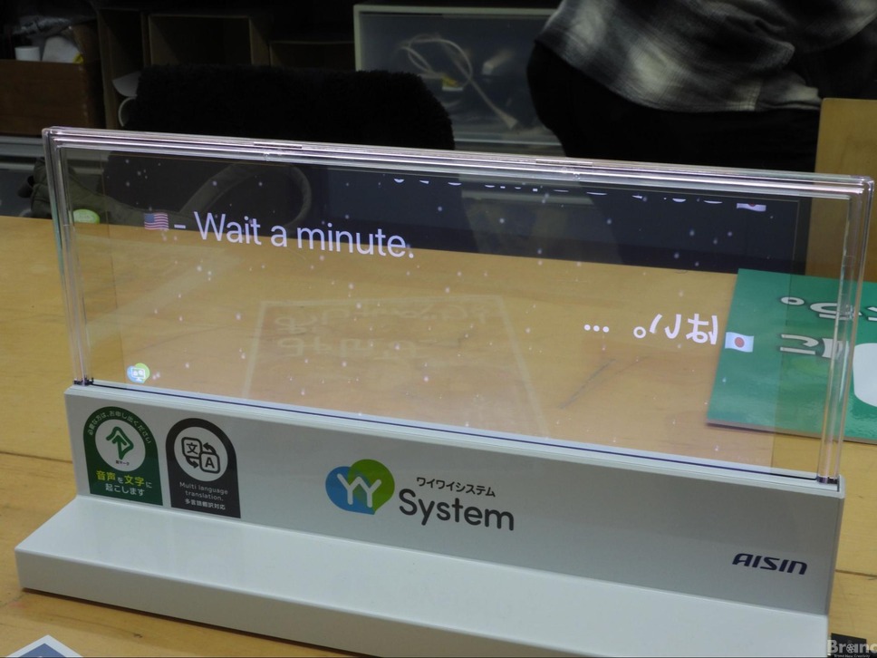 机の上に置かれた、アイシンの透明ディスプレイ搭載の翻訳・字幕表示ツール「YY System（ワイワイシステム）」。向こう側が透けて見えるスクリーンに、アメリカ国旗のアイコンと「Wait a minute.」という文字が表示されています。本体には「音声を文字に起こします」「多言語翻訳対応」という案内ステッカーが貼られている。