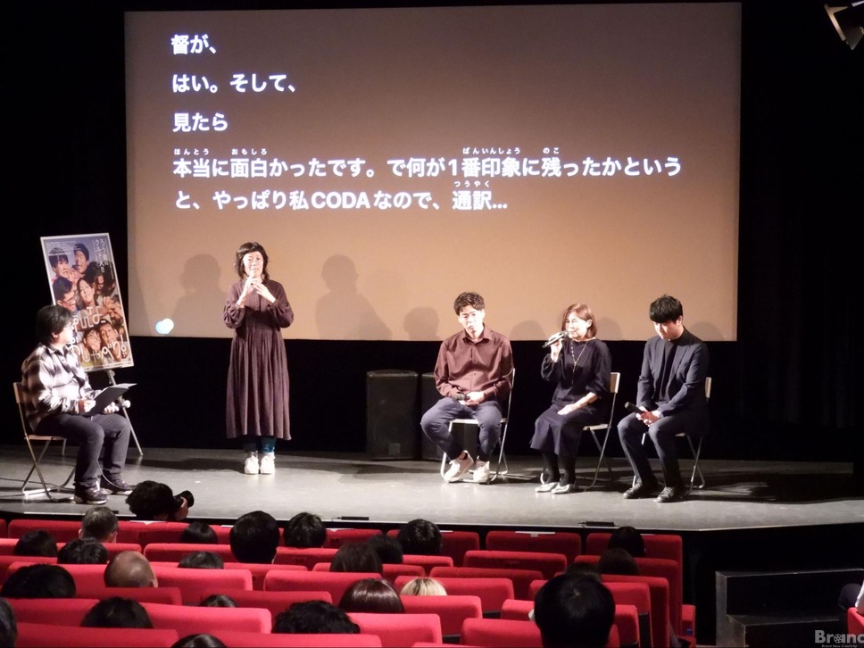 映画館のステージで行われているトークイベントの様子。ステージ背景の巨大なスクリーンには、登壇者の発言が「...やっぱり私CODAなので...」と、読み仮名付きの大きな白い文字で字幕表示されている。ステージ上には、手話通訳を行う女性が立っており、その横でマイクを持った女性を含む3名のゲストが座って話をしている。