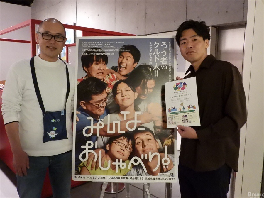 映画『みんなのおしゃべり！』の大型ポスターを間に挟んで並ぶ2人の男性の記念写真。左側は株式会社アイシンの中村正樹氏で、同社の「YY System」のロゴ入りポーチを首から下げている。右側は映画の河合健監督で、手にチラシを持っている。中央のポスターには「ろう者 vs クルド人!!」「通じ合わない人たちが、大混戦!!」などのキャッチコピーと共に、キャストたちの賑やかな表情がデザインされている。