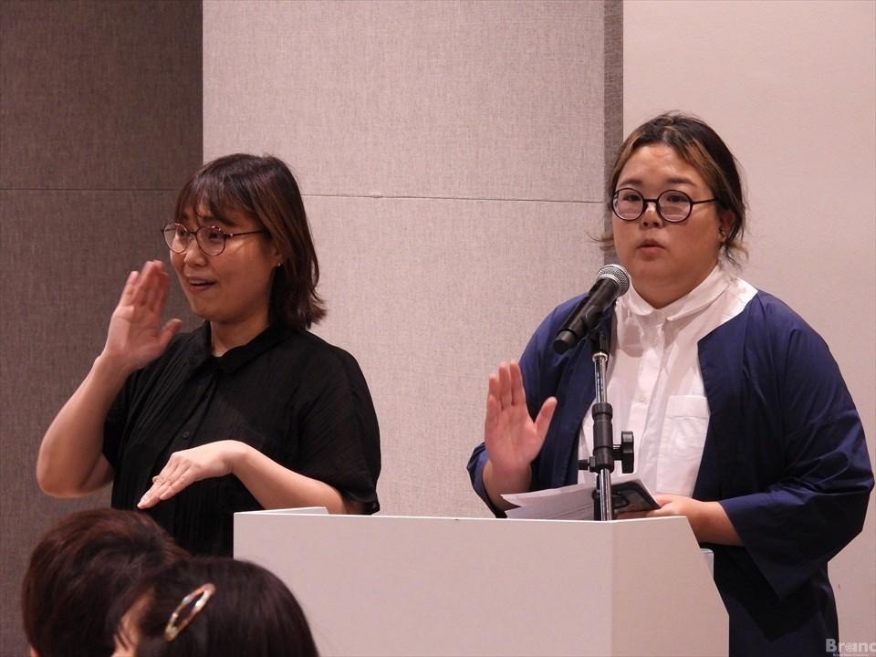 白い演台に立つ2人の人物。右側の人物はマイクに向かって話し、左側の人物は手話を行っている