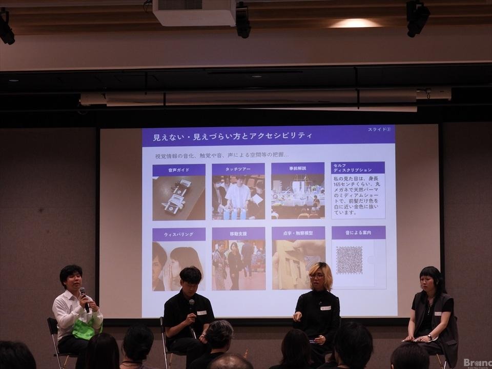 4人の登壇者が並んで座るパネルディスカッションの様子。背後のスクリーンには『見えない・見えづらい方とアクセシビリティ』と題されたスライドが投影され、音声ガイドなどの事例が写真付きで紹介されている