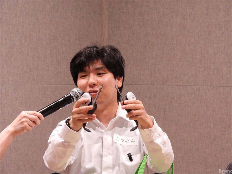 『たつみん』という名札をつけた男性の鈴木龍弥さんが、両手に黒いあしらせを持ち、差し出されたマイクに向かって笑顔で話している様子